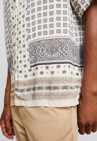 Lyhythihainen nappipaita vaaleassa kermavärissä, jossa on sinisiä ja ruskeita monimutkaisia kuvioita. Paidassa on rento malli ja tekstuurilla varustettu kangas.