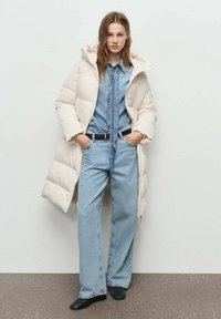 Abrigo largo acolchado en beige claro, combinado con una camisa de mezclilla azul y jeans de pierna ancha. Un cinturón negro y botas completan el conjunto.