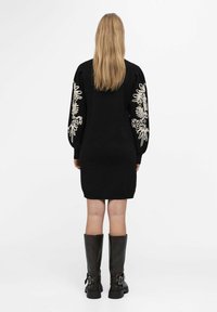 Femme aux longs cheveux blonds portant une robe noire avec une broderie florale blanche sur les manches et des bottes noires montantes, debout de dos.