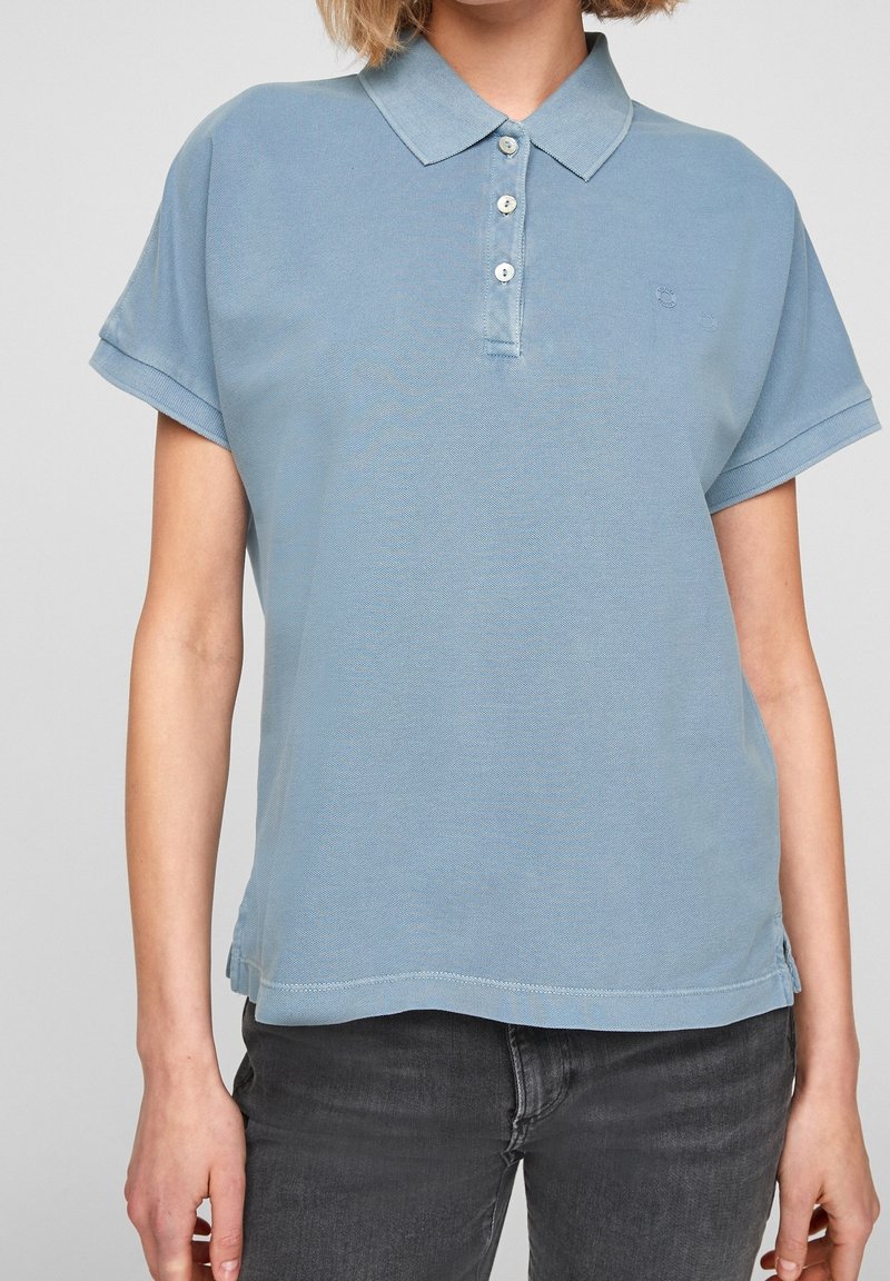 Vaaleansininen polo-paita, jossa on kaulus, kolmen napin nappilista, lyhyet hihat ja hienovarainen logo brodeerattuna rintaan. Puuvillasekoitekangas.