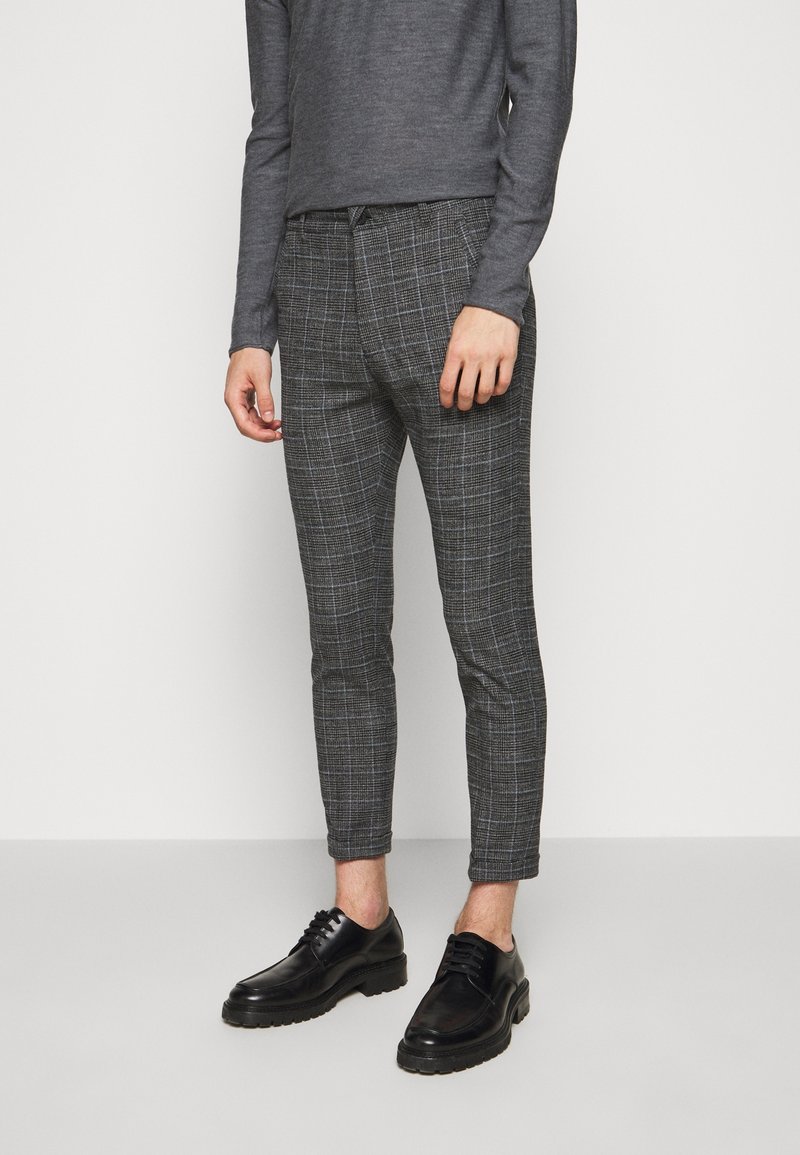 Homme portant un pantalon gris à carreaux ajusté, un haut à manches longues gris foncé et des chaussures noires à lacets, debout sur fond blanc.