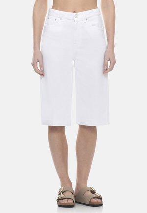 Donna che indossa pantaloncini di jeans bianchi fino al ginocchio e sandali beige con doppia fascia, in piedi con le braccia rilassate ai lati.