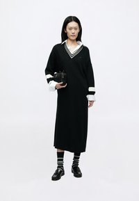 Črna oversized obleka z belimi črtastimi ovratnikom in manšetami, kombinirana s črnimi čevlji in črtastimi nogavicami, v rokah pa manjša črna torbica.