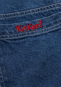 Tessuto denim di colore blu scuro con texture evidente. Logo ricamato rosso "Kickers" sul bordo della tasca posteriore, con cuciture bianche a contrasto.