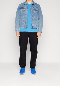 Giacca in denim azzurro sopra un maglione blu, abbinato a pantaloni neri e sneakers blu brillante. Look casual e stratificato.