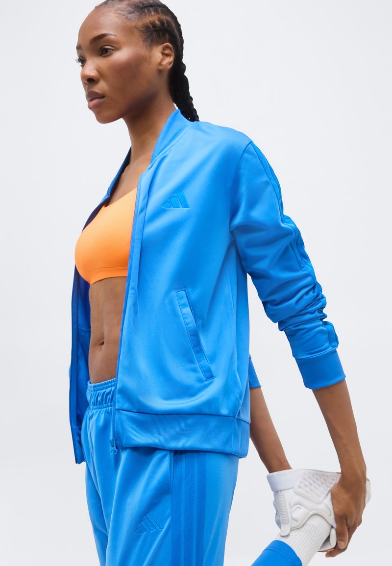 Donna in tuta sportiva blu e reggiseno sportivo arancione che si allunga tenendo una scarpa da ginnastica bianca dietro la schiena.