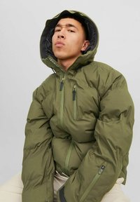 Chaqueta verde acolchada con capucha, que cuenta con dos bolsillos en el pecho con cremallera y un detalle de logo. Tejido texturizado con acabados elásticos en las muñecas.