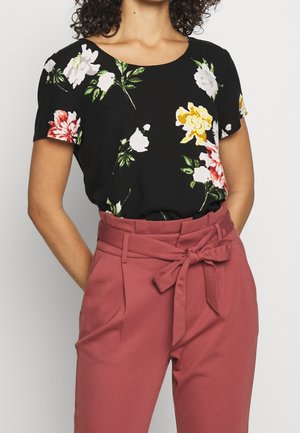 Frau trägt ein schwarzes florales Kurzarmoberteil, das in hoch taillierte mauvefarbene Hose mit einem gebundenen Gürtel an der Taille gesteckt ist.