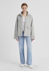 Graue Wollmischungjacke mit hohem Kragen, Vorderreißverschluss und zwei aufgesetzten Taschen. Lockere Passform mit weiten Ärmeln, getragen über einem weißen Oberteil und Jeans.