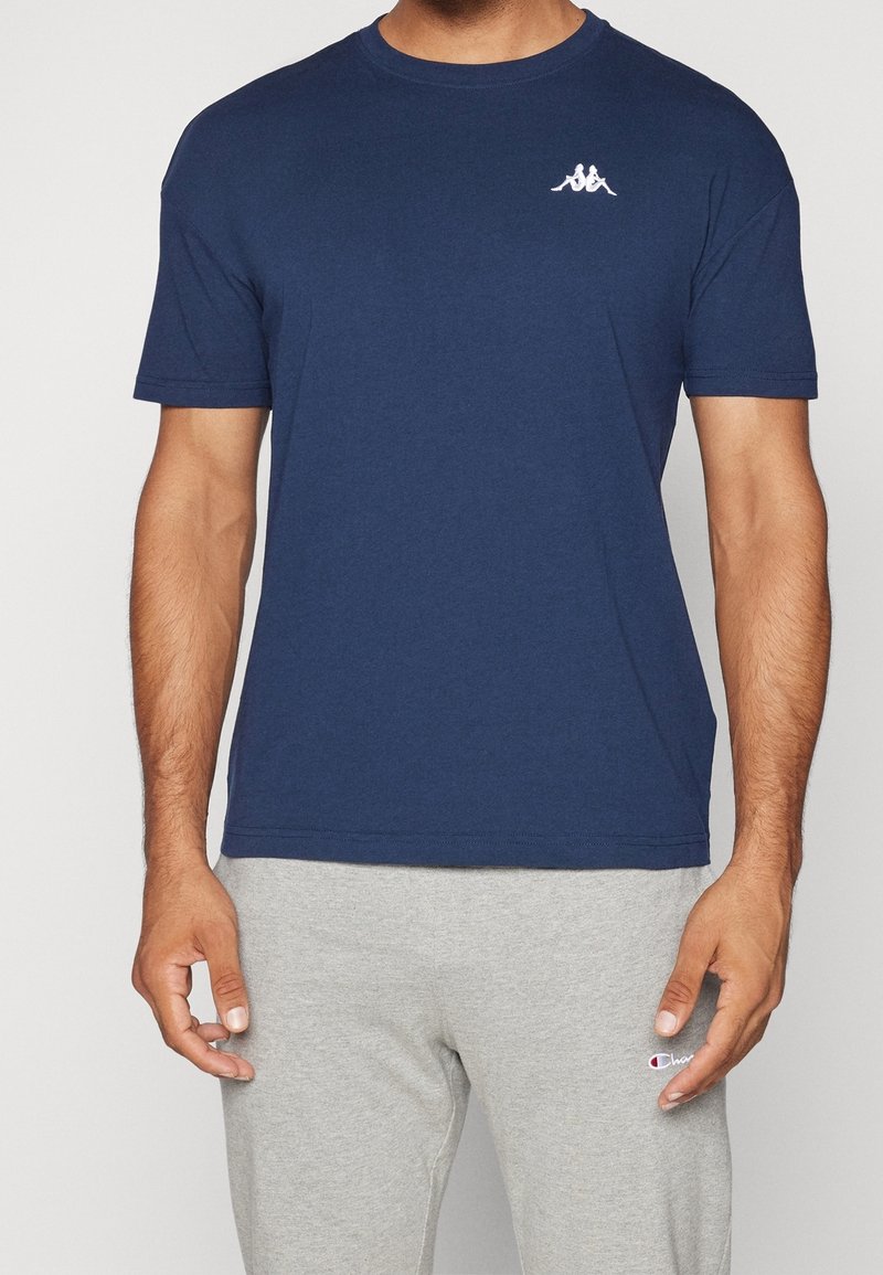 Homme portant un t-shirt bleu marine avec un petit logo blanc et un pantalon de jogging gris clair avec un petit logo sur la cuisse.