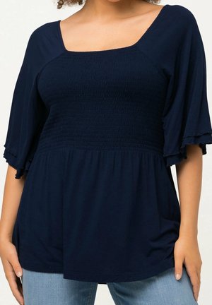 Vrouw die een marineblauw ingesnoerd topje met vierkante hals en vlindermouwen tot aan de ellebogen draagt, gecombineerd met lichtblauwe jeans, staand met ontspannen armen.