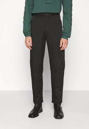 Homme portant un pantalon noir droit, des bottines en cuir noir et une chemise verte à manches longues avec des détails aux manches, debout devant un fond blanc.