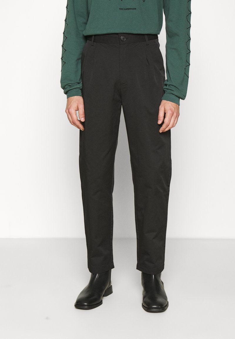 Homme portant un pantalon noir droit, des bottines en cuir noir et une chemise verte à manches longues avec des détails aux manches, debout devant un fond blanc.