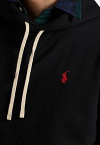 Felpa nera con cappuccio e logo ricamato rosso, cordini crema, sovrapposta a una camicia a quadri blu e verde. Tessuto morbido e design casual.