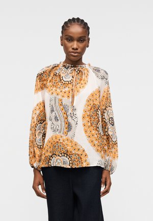 Frau mit geflochtenem Haar, die eine langärmlige Bluse mit orange-schwarzen und weißen kreisförmigen Blumenmustern sowie eine schwarze Hose trägt.