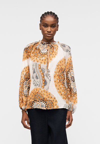 Femme aux cheveux tressés portant un chemisier à manches longues avec des motifs floraux circulaires orange, noir et blanc, ainsi qu'un pantalon noir.