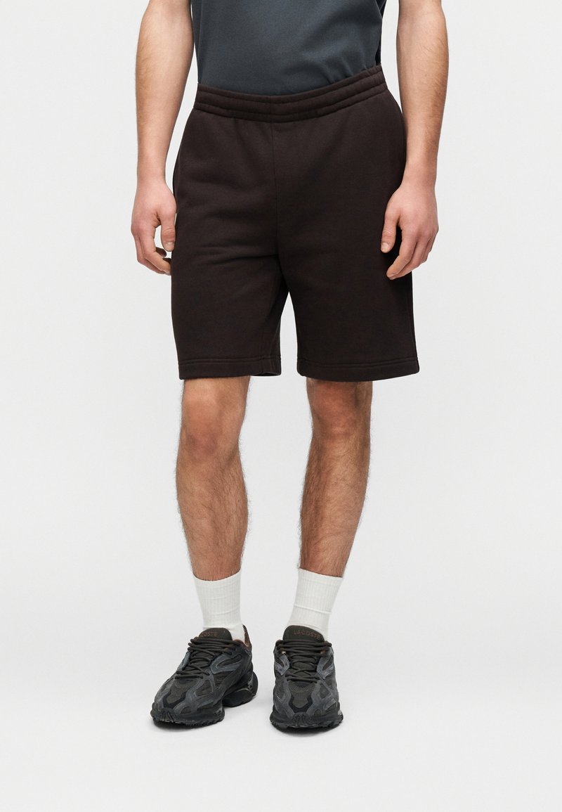 Shorts en coton marron foncé avec une taille élastique, dotés d'une coupe classique. Portés avec des chaussettes blanches mi-mollet et des baskets noires à motifs.