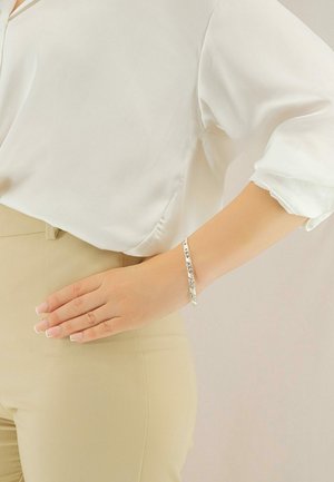 Bracciale in argento con maglie, indossato su un polso. Lo sfondo presenta un colore grigio chiaro e il polso ha una finitura neutra.