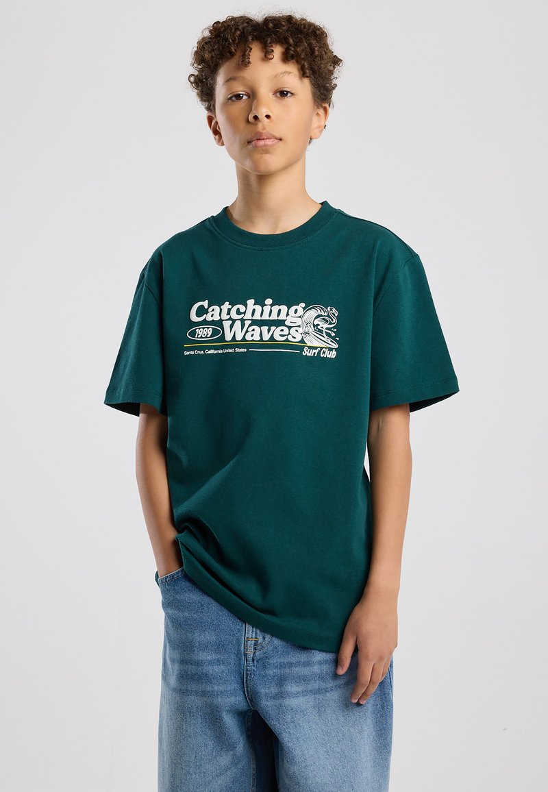 Niño con cabello rizado que lleva una camiseta verde oscuro con el texto "Catching Waves Surf Club" y pantalones vaqueros azules, de pie con una mano en el bolsillo.