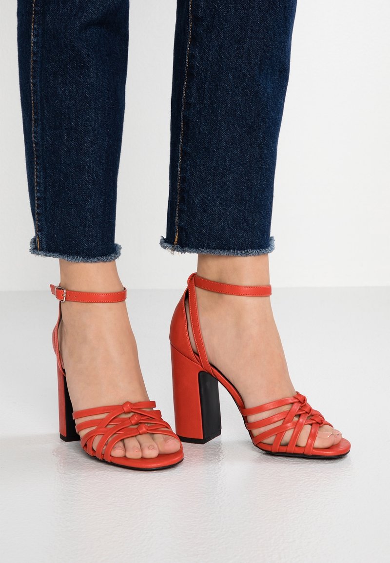 Pieds portant des sandales rouges à talons hauts avec brides à la cheville, associées à un jean bleu foncé raccourci avec des ourlets effilochés.