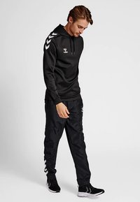 Hoodie de sport noir avec une poche avant, arborant un logo blanc et des bandes en forme de chevrons sur les manches ; assorti à un pantalon de survêtement noir et des chaussures sportives.