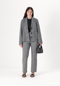 Grå og svart rutete blazer med to lommer, parret med matchende bukser og en svart håndveske. Modellen har på seg svart topp og sandaler med stropper.