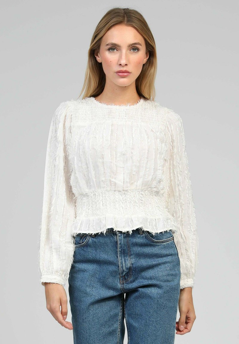 Femme aux cheveux blonds raides portant un blouse blanche texturée à manches longues et un jean bleu taille haute, se tenant devant un fond uni.