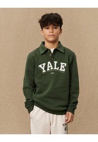 Grønn sweatshirt med polo-krage, med "YALE" i hvite bokstaver. Laget av myk stoff, med ribbede mansjetter og kant. Kombinert med kremfargede bukser.