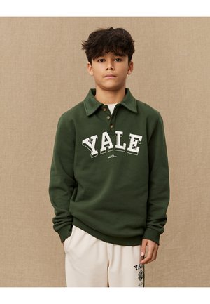 Grønn sweatshirt med polo-krage, med "YALE" i hvite bokstaver. Laget av myk stoff, med ribbede mansjetter og kant. Kombinert med kremfargede bukser.