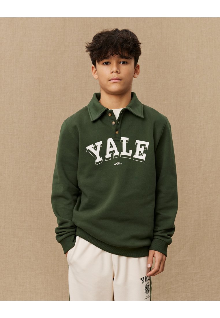 Grønn sweatshirt med polo-krage, med "YALE" i hvite bokstaver. Laget av myk stoff, med ribbede mansjetter og kant. Kombinert med kremfargede bukser.