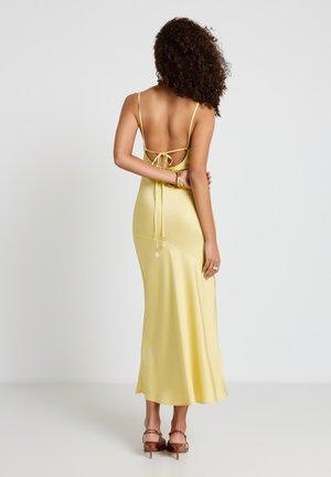 Femme aux cheveux bouclés portant une robe midi jaune en satin sans dos avec des bretelles spaghetti et des sandales à talons marron, debout devant un fond blanc.