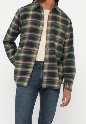 Mężczyzna w zielono-brązowo-białej koszuli w kratę zapinanej na guziki, na beżowej bluzce, w połączeniu z niebieskimi dżinsami, stojący na białym tle.