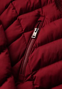Bordowy puchowy płaszcz z pikowaną fakturą, wyraźnym zamkiem błyskawicznym z srebrnymi elementami i gładkim wykończeniem.