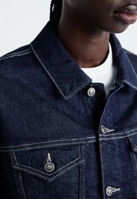 Mörk denimjacka med klassisk krage, silverfärgad knappraring och kontrasterande vit sömnad. Har bröstfickor och strukturerad tyg.