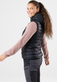 Svart puffärmad väst med huva, med ett kviltat mönster. Bärs över en ljusrosa fleece-tröja, modellen poserar med en lätt vridning som visar sidodetaljer.