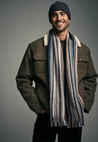 Brown wool-blend jacket with a cream shearling collar, paired with a multicolored striped scarf featuring blue, beige, and white tones.