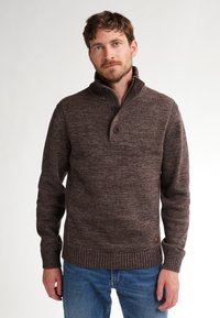 Man met een bruine gebuttonde gebreide trui met geribde manchetten en zoom, gecombineerd met blauwe jeans, staande tegen een effen witte achtergrond.