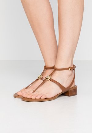 Sandalias T-bar de cuero marrón con detalles de herrajes en tono dorado, tacón plano y diseño de punta abierta que exhibe uñas de los pies pulidas.