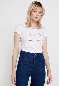 Baltas medvilnės marškinėlis su užrašu "A|X ARMANI EXCHANGE" iš nertų rašmenų, derinamas su aukšto juosmens tamsiomis džinsomis su priekinių kišenių.