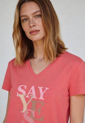 Junge Frau mit schulterlangem hellbraunem Haar, die ein korallenfarbenes V-Ausschnitt-T-Shirt mit dem Text „SAY YES“ in gemischten Schriftarten trägt.