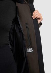 Giacca con tessuto esterno nero e interno marrone. Presenta una tasca con cerniera e un'etichetta che recita "Khujo Original Changing Threads."
