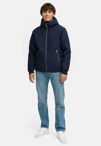 Marinejakke med hette, glidelås foran, to sidelommer, ribbestrikkede ermer, brukt over lyseblå jeans og hvite sneakers.