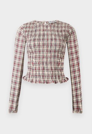 Ilgi piedurkņu, pieguļoša rūtainā blūze ar nazumotu audumu un volānu apakšmalu bēša, sarkanu un zilu toņu kombinācijā, attēlota uz vienkārša fona.