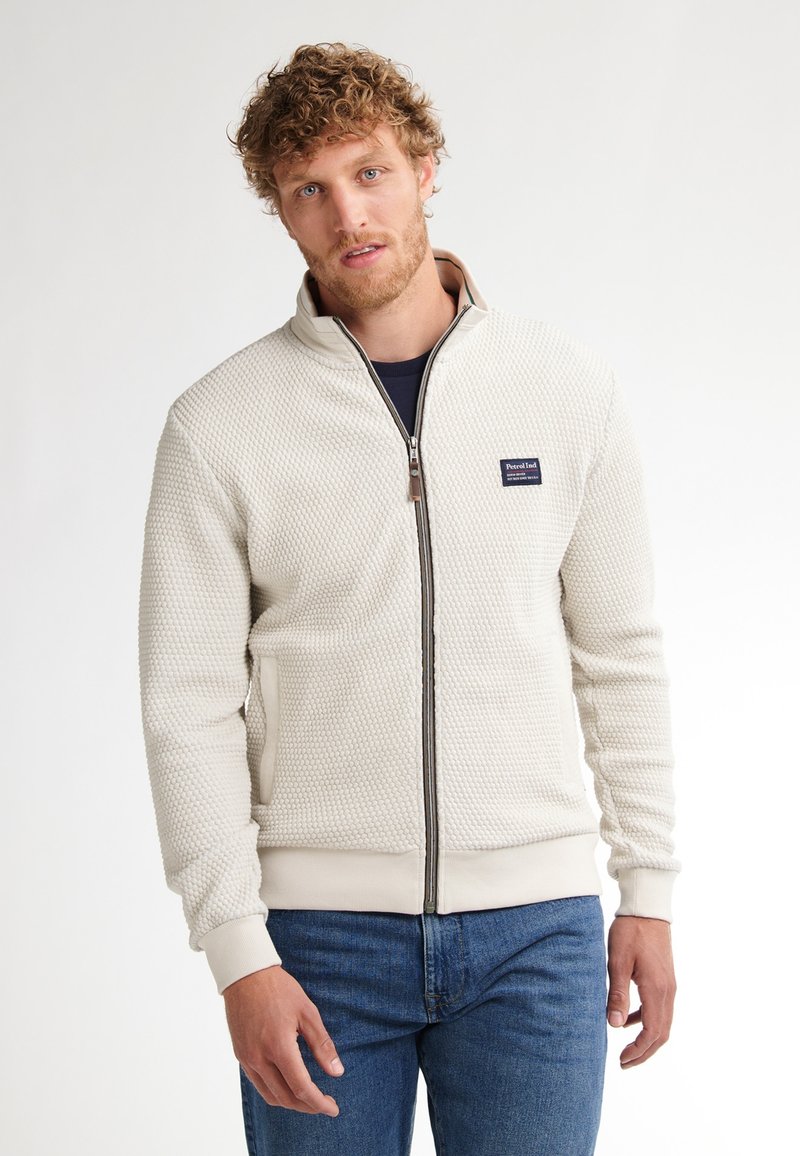 Texturovaná krémová zipová bunda s vysokým límcem, obsahující malou nášivku loga na hrudi a kontrastní vroubkované manžety a lem.