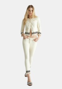 Chaqueta de marfil corta con dobladillo de flecos, combinada con pantalones a juego que presentan flecos y acentos texturizados. Modelo con sandalias decorativas.