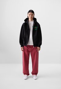 Black zip-up hoodie with green graphic, layered over a white t-shirt, paired with loose red pants and white sneakers. Simple, casual attire.