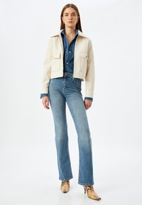Cremfarbener cropped Blazer mit zwei frontalen Taschen. Darunter ein blaues Denim-Hemd. Flared Blue Jeans und beigefarbene Schlangen-muster Stiefeletten mit Absatz runden das Outfit ab.