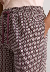 Close-up van gebloemde broek met bordeauxrode en witte geometrische vormen, elastische tailleband, trekkoord en een hand in de rechterzak.