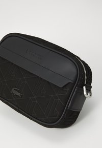 Borsa Lacoste nera con una parte frontale in tessuto texturizzato con motivi geometrici, una parte superiore liscia e una chiusura con zip silver.