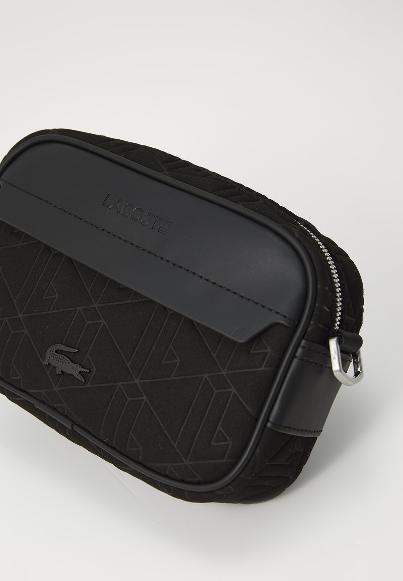 Sac Lacoste noir avec un devant en tissu texturé aux motifs géométriques, un dessus lisse et une fermeture à glissière en argent.